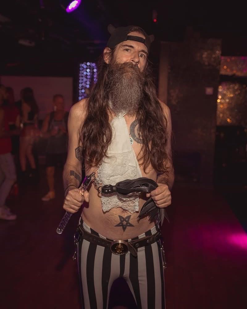 MR. SATAN holding a riding crop.